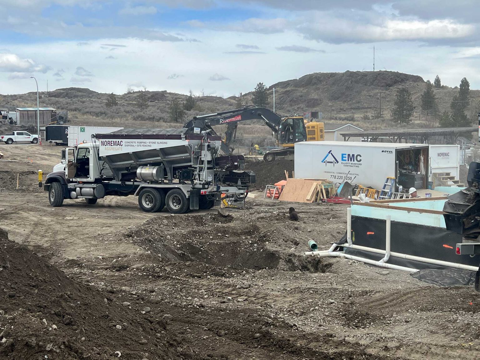 A Noremac stone slinger on site with the words Noremac Aggregate on it, an EMC trailer with other construction equipment in the background.