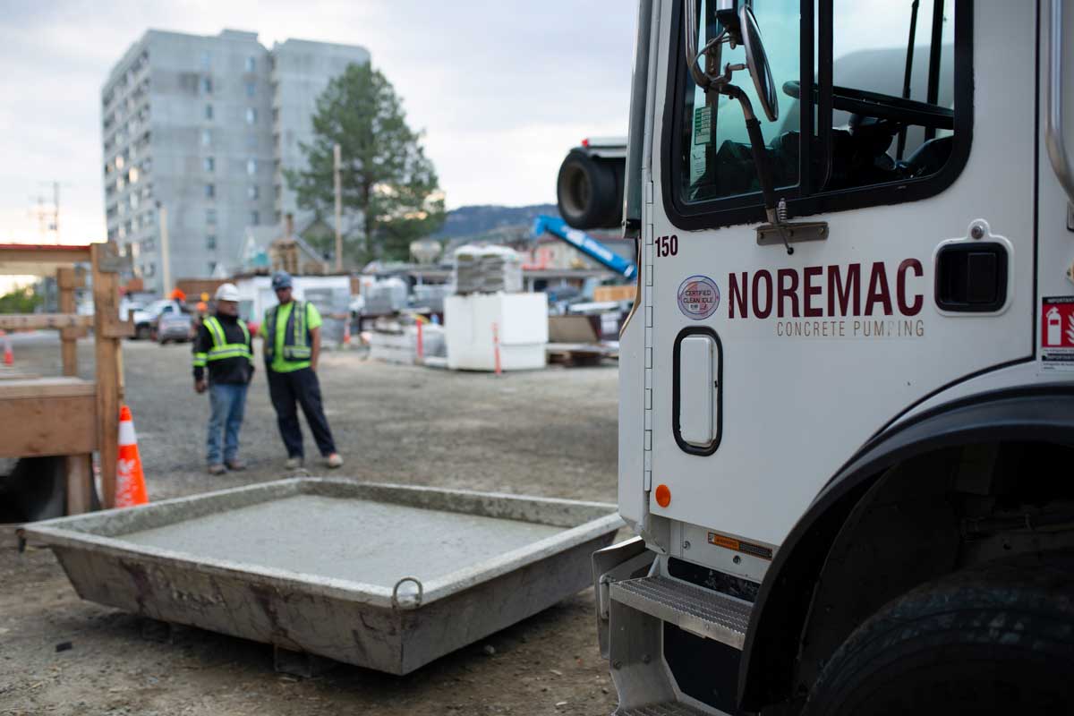 A Noremac-branded concrete pumping truck, concrete in a container in the background of a construction site.
