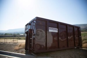 A burgundy Noremac disposal bin, with a blue sunny sky in the background.