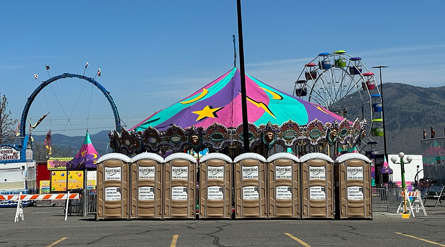 Noremac portable toilets in a row at a fair ground, a carousel, roller coaster, and ferris wheel in the background.