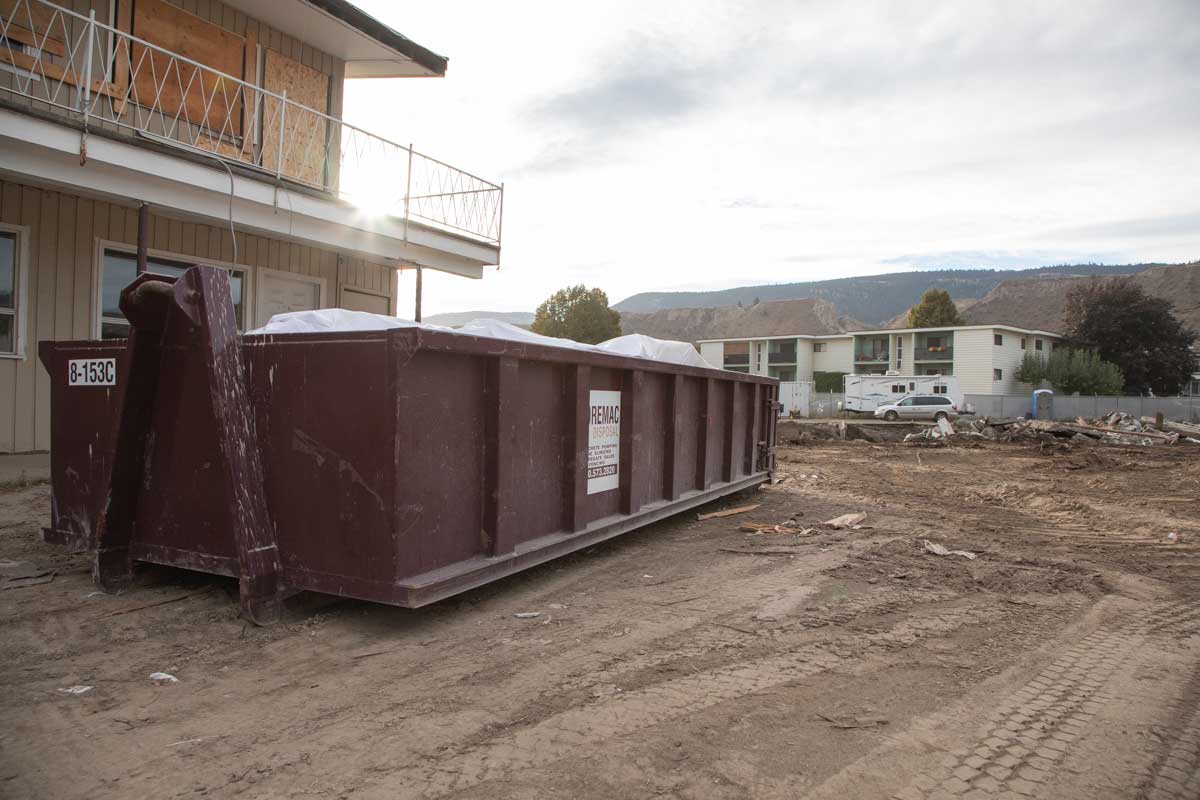 A construction site for a residential condo, with a large maroon bin in front, the Noremac Disposal logo with the Noremac phone number on a sign. The Noremac phone number is 250-573-2820.