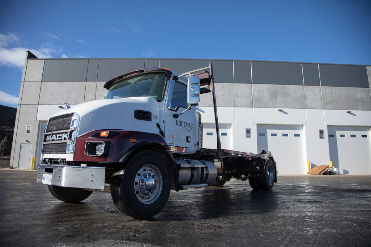 A Noremac roll-off bin truck is poised in front of an industrial storage warehouse, garage doors in the background.