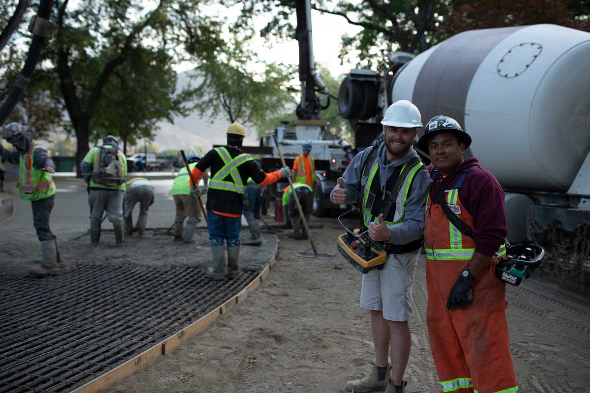 Two concrete pumping workers smile at the camera. One of them is giving two big thumbs up, a concrete pouring truck in the background, with the rest of the team working hard pouring concrete.